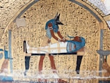 An ancient Egyptian art piece depicting Anubis, the jackal-headed god, performing a ritual on a mummified figure. Hieroglyphics fill the background, adding to the historical and cultural context.