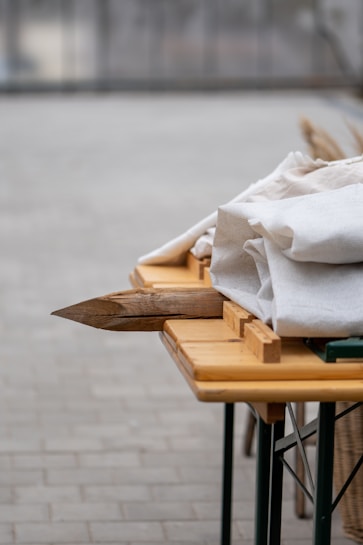 A wooden table with a bundle of light-colored fabric on top, set outdoors on a paved surface. Wooden stakes are visible, adding a rustic element to the composition. The background is blurred, emphasizing the focus on the table and objects.