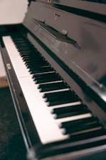 A close-up view of a black upright piano, focusing on the keyboard. The keys are alternately black and white, with a reflective surface showing some of the surroundings. The piano's glossy finish adds a touch of elegance.