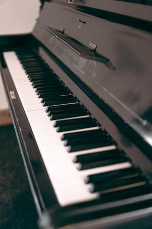 A close-up view of a black upright piano, focusing on the keyboard. The keys are alternately black and white, with a reflective surface showing some of the surroundings. The piano's glossy finish adds a touch of elegance.