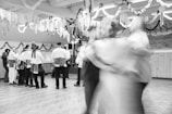 A group of people dressed in traditional attire are gathered in a decorated indoor space, possibly for a cultural or festive event. The room is adorned with streamers and paper decorations hanging from the ceiling. In the foreground, a couple is dancing in motion, creating a blurred effect.
