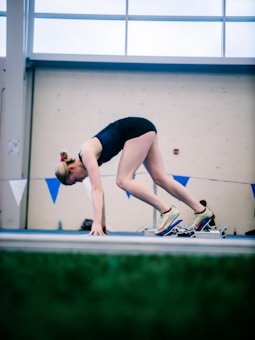 A person is in a crouched starting position on a track, preparing for a sprint or race. They wear athletic clothing and running shoes, with focus and readiness in their posture. Blue and white triangular flags are visible in the background, with part of a glass window letting in natural light.