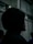 A person wearing a dark hooded garment is standing in a dimly lit environment with blurred backgrounds, creating a mysterious and moody atmosphere.