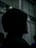 A person wearing a dark hooded garment is standing in a dimly lit environment with blurred backgrounds, creating a mysterious and moody atmosphere.