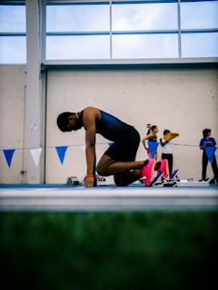 An athlete in a blue uniform is crouching at the starting blocks on an indoor track, preparing for a race. The focus is on the runner's vibrant pink shoes. In the background, other athletes and officials are visible, suggesting a competitive event. The setting appears to be a large, well-lit indoor sports facility.