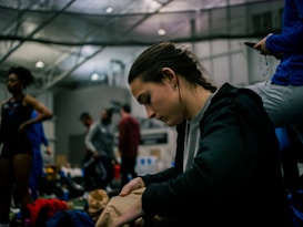 Several people are inside a large indoor facility. The focus is on an individual in the foreground who is deeply concentrated, possibly packing or unpacking an item. Other persons are visible in the background, some standing and engaging in various activities.