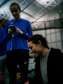 Two people are indoors in what appears to be a sports facility with a high ceiling and metal framework. One person is looking down at their phone with earphones in, wearing a blue long-sleeve athletic shirt. The other person is smiling and appears to be engaging with something or someone off-camera, dressed in darker athletic wear. The background is slightly blurred but shows additional people and possibly sports equipment.