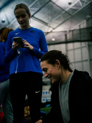 Two people are indoors in what appears to be a sports facility with a high ceiling and metal framework. One person is looking down at their phone with earphones in, wearing a blue long-sleeve athletic shirt. The other person is smiling and appears to be engaging with something or someone off-camera, dressed in darker athletic wear. The background is slightly blurred but shows additional people and possibly sports equipment.