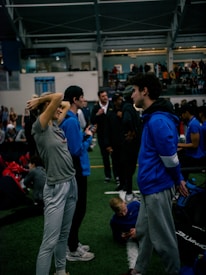 People engage in conversation and activities in an indoor sports facility. A woman in gray athletic wear stretches her arms above her head while talking to a man in a blue jacket. Other people are seen interacting in the background, some are seated on the ground, and the atmosphere is lively with many individuals gathered.