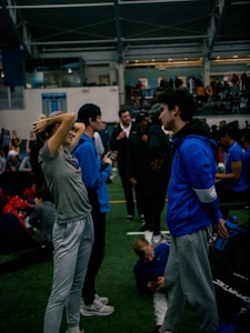 People engage in conversation and activities in an indoor sports facility. A woman in gray athletic wear stretches her arms above her head while talking to a man in a blue jacket. Other people are seen interacting in the background, some are seated on the ground, and the atmosphere is lively with many individuals gathered.