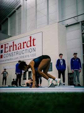 An athlete in a sprint starting position on an indoor track, surrounded by several people who seem to be spectators or fellow athletes. The area has a large construction sign on the wall in the background, and the setting looks like an indoor sports facility.