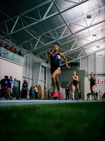 Athletes are competing in a relay race inside a large sports facility. The runners are moving swiftly along the track, with one runner in the foreground captured mid-stride. Spectators and other participants can be seen in the background, adding to the dynamic and energetic atmosphere of the scene.