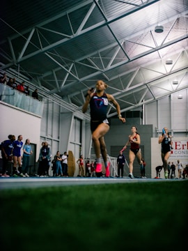 Athletes are competing in a relay race inside a large sports facility. The runners are moving swiftly along the track, with one runner in the foreground captured mid-stride. Spectators and other participants can be seen in the background, adding to the dynamic and energetic atmosphere of the scene.