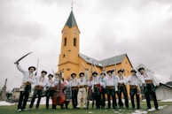 A group of people dressed in traditional attire stand in front of a yellow church building with a tall spire. Many of them are holding swords, and one person is holding a stringed instrument similar to a double bass. The sky is overcast, giving a muted backdrop to the vibrant clothing and building.