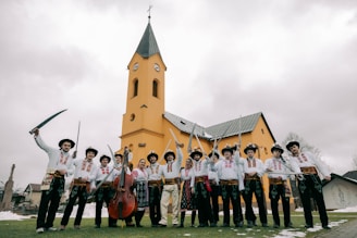 A group of people dressed in traditional attire stand in front of a yellow church building with a tall spire. Many of them are holding swords, and one person is holding a stringed instrument similar to a double bass. The sky is overcast, giving a muted backdrop to the vibrant clothing and building.