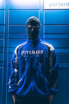 A person wearing a blue Yamaha racing jacket and a black balaclava mask stands in front of a background with blue lockers.