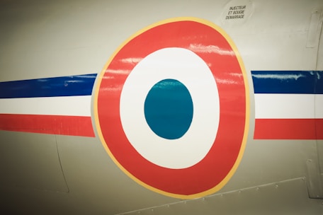 A close-up view of a circular emblem painted on the side of an aircraft. The emblem consists of concentric circles in red, white, and blue, resembling markings often found on military or vintage aircraft. The surface displays a smooth metal texture, with rivets visible along the edges.
