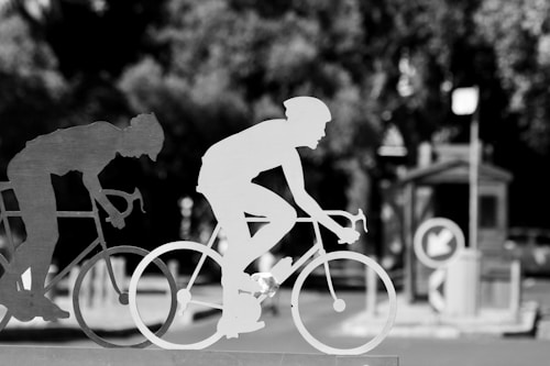 Metallic silhouettes of cyclists are prominently displayed in the foreground, with one positioned slightly ahead of the other, conveying a sense of motion. In the background, elements of an urban environment, such as a street sign and a small structure, are slightly out of focus.