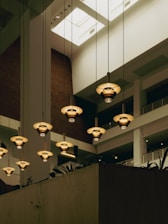 A modern interior setting with multiple hanging pendant lights. The lights have a warm, golden hue and are suspended from the high ceiling. The architecture features clean lines, brick walls, and large open spaces with a skylight allowing natural light to enter. Indoor plants are visible on the lower ledge.