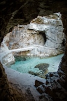 A natural rock formation resembling a cave with a small pool of clear, turquoise water inside. The cave's walls are rough and jagged, made of light and dark stone layers. Sunlight filters in, highlighting the serene and tranquil setting. A narrow concrete pathway extends over part of the water.