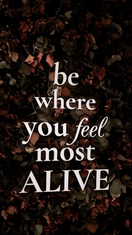 Bold, white text with the motivational message 'be where you feel most ALIVE' is overlaying a background of dark, leafy foliage. The leaves are predominantly dark green with earthy brown and muted red tones, contributing to a natural and organic look.
