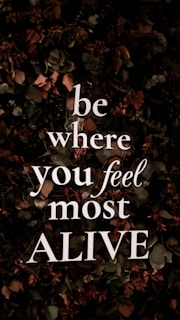 Bold, white text with the motivational message 'be where you feel most ALIVE' is overlaying a background of dark, leafy foliage. The leaves are predominantly dark green with earthy brown and muted red tones, contributing to a natural and organic look.