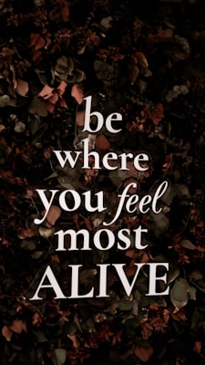 Bold, white text with the motivational message 'be where you feel most ALIVE' is overlaying a background of dark, leafy foliage. The leaves are predominantly dark green with earthy brown and muted red tones, contributing to a natural and organic look.