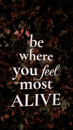 Bold, white text with the motivational message 'be where you feel most ALIVE' is overlaying a background of dark, leafy foliage. The leaves are predominantly dark green with earthy brown and muted red tones, contributing to a natural and organic look.