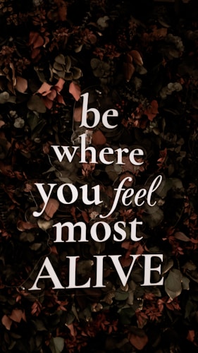 Bold, white text with the motivational message 'be where you feel most ALIVE' is overlaying a background of dark, leafy foliage. The leaves are predominantly dark green with earthy brown and muted red tones, contributing to a natural and organic look.