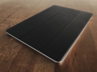 A sleek, dark-colored tablet sits on a wooden surface, with light reflecting off the smooth tablet cover. The wood has a warm tone, and the tablet is positioned at a slight angle.