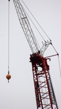 A large construction crane with a red metal framework and a long white lattice boom is raising a heavy-duty hook. The background is overcast with a gray sky.