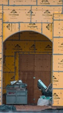 A construction site with an archway framed in DensGlass sheathing panels. There is a large plywood board partially covering the entrance. Construction materials, including a metal container and drywall, are scattered around.