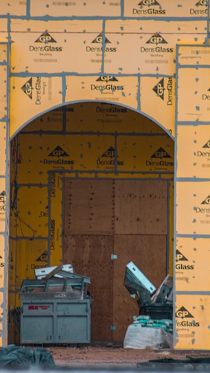 A construction site with an archway framed in DensGlass sheathing panels. There is a large plywood board partially covering the entrance. Construction materials, including a metal container and drywall, are scattered around.