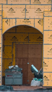 A construction site with an archway framed in DensGlass sheathing panels. There is a large plywood board partially covering the entrance. Construction materials, including a metal container and drywall, are scattered around.