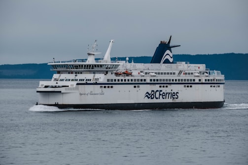 A large ferry named 'Spirit of Vancouver Island' is sailing on a calm body of water. The ferry is part of the BC Ferries fleet and is predominantly white with blue accents. The background features a hazy, distant coastline with forested hills under a gray sky.