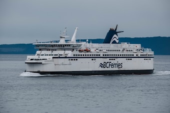 A large ferry named 'Spirit of Vancouver Island' is sailing on a calm body of water. The ferry is part of the BC Ferries fleet and is predominantly white with blue accents. The background features a hazy, distant coastline with forested hills under a gray sky.