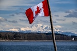 A Canadian flag is prominently displayed in the foreground, waving against a backdrop of snow-capped mountains. A serene body of water lies between the viewer and a forested area, leading into the majestic mountain range under a partly cloudy sky.