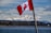 A Canadian flag is prominently displayed in the foreground, waving against a backdrop of snow-capped mountains. A serene body of water lies between the viewer and a forested area, leading into the majestic mountain range under a partly cloudy sky.