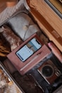 An open suitcase filled with neatly packed clothes, a smartphone displaying a banking app, a notebook with a pen on top, a digital camera, and sunglasses. The suitcase is organized and ready for travel.