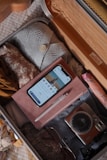 An open suitcase filled with neatly packed clothes, a smartphone displaying a banking app, a notebook with a pen on top, a digital camera, and sunglasses. The suitcase is organized and ready for travel.