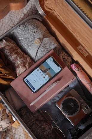 An open suitcase filled with neatly packed clothes, a smartphone displaying a banking app, a notebook with a pen on top, a digital camera, and sunglasses. The suitcase is organized and ready for travel.