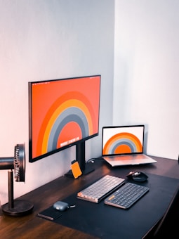 An organized workspace featuring a large computer monitor and an open laptop displaying identical wallpapers with a colorful, concentric circle design. The desk also has a keyboard, mouse, smartphone, and a phone holder next to the monitor.