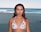 A person stands on a sandy beach with the ocean behind them. They are wearing a white bikini top with a cherry pattern, accessorized with earrings and a necklace. The ocean has gentle waves, and the sky is clear, suggesting a calm, sunny day.