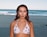 A person stands on a sandy beach with the ocean behind them. They are wearing a white bikini top with a cherry pattern, accessorized with earrings and a necklace. The ocean has gentle waves, and the sky is clear, suggesting a calm, sunny day.