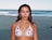A person stands on a sandy beach with the ocean behind them. They are wearing a white bikini top with a cherry pattern, accessorized with earrings and a necklace. The ocean has gentle waves, and the sky is clear, suggesting a calm, sunny day.