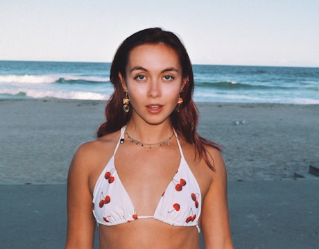 A person stands on a sandy beach with the ocean behind them. They are wearing a white bikini top with a cherry pattern, accessorized with earrings and a necklace. The ocean has gentle waves, and the sky is clear, suggesting a calm, sunny day.