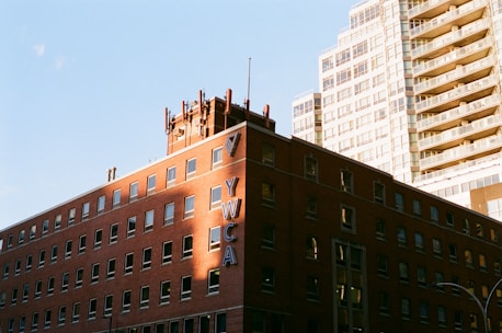 A brick building with multiple windows displays the large vertical letters 'YWCA' on the side. The building is bathed in sunlight, casting warm shadows. In the background, a high-rise building with a modern glass facade rises against a clear blue sky.