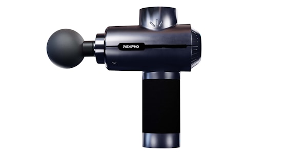 A handheld massage gun with a sleek, modern design featuring a spherical massage head attached to a cylindrical body. The gun is primarily black with metallic accents and displays a logo on its side.