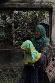 A person wearing a green hijab and a plaid outfit is holding a child dressed in a vibrant green and yellow headscarf against a backdrop of a weathered wall and metal grid with lush greenery.