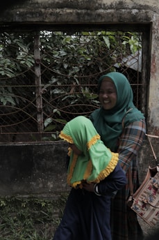 A person wearing a green hijab and a plaid outfit is holding a child dressed in a vibrant green and yellow headscarf against a backdrop of a weathered wall and metal grid with lush greenery.